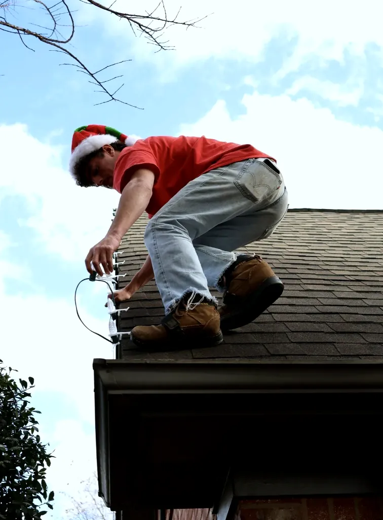 Installer crouched on a roof above the gutter, clipping a light strand in place.