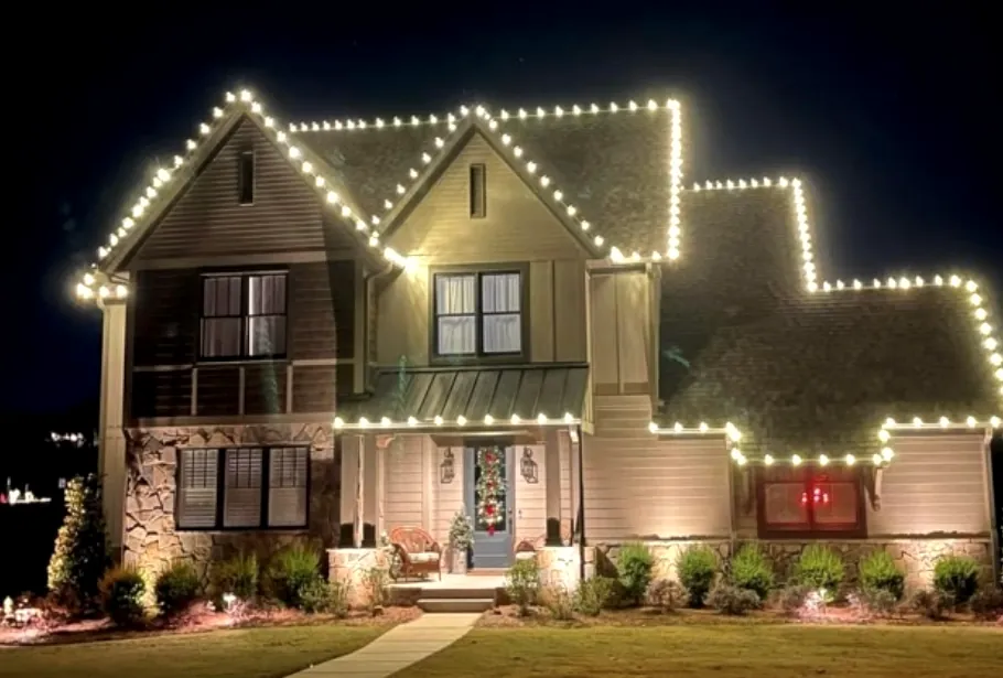 Multi-gable home with a lit roofline and warm uplights across the garden beds.