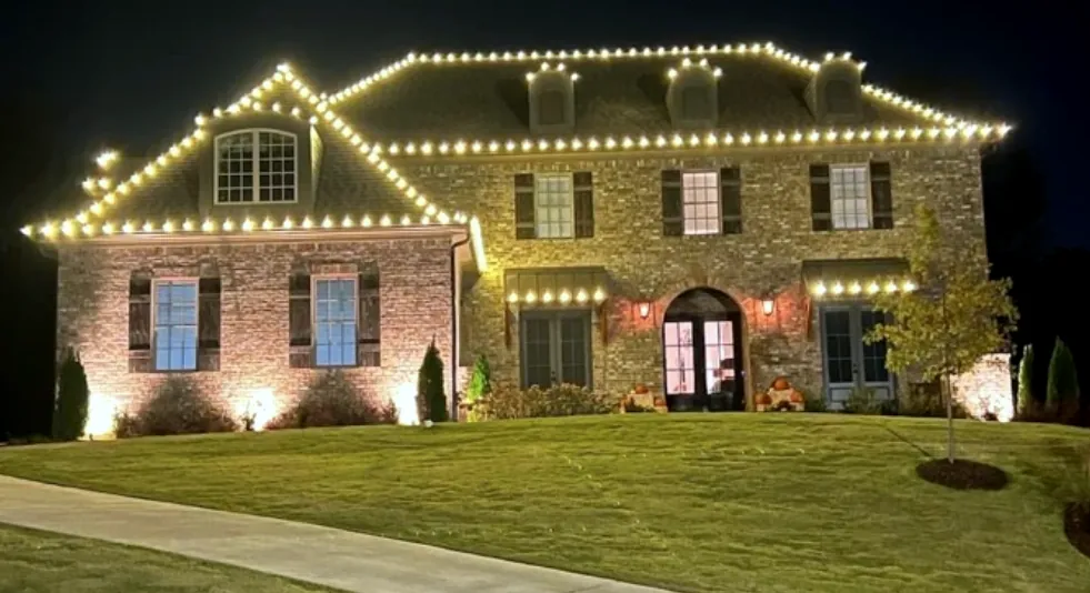 Stone and brick two-gable house with a crisp white roofline above a wide front lawn at night.