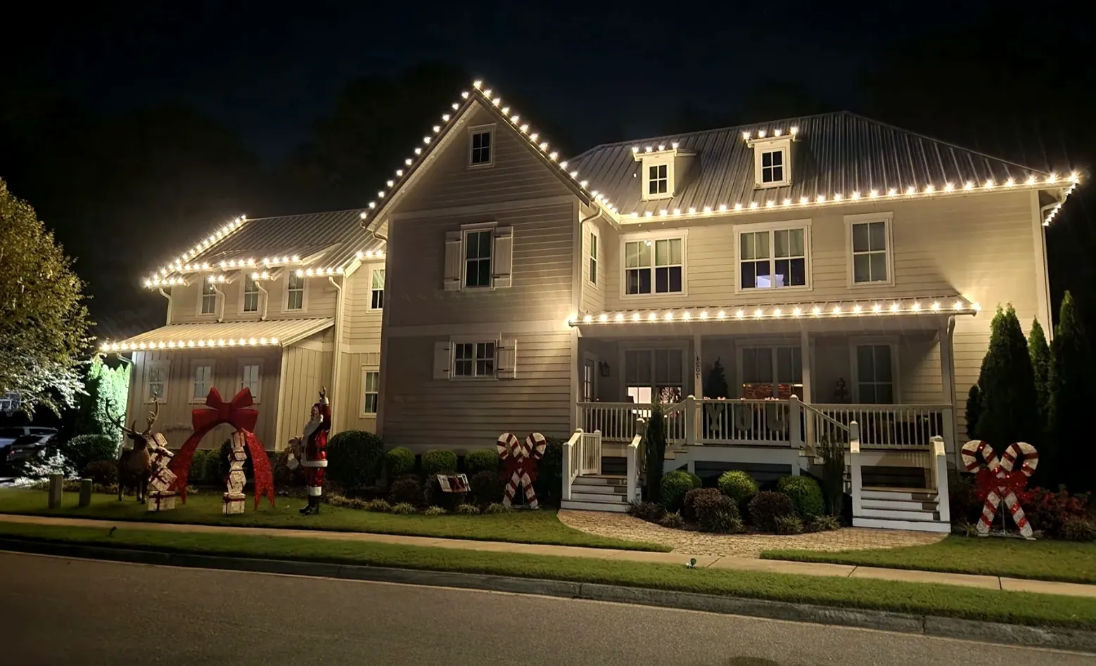 Two-story gray house with a covered porch, red bows and lights tracing every roof edge.